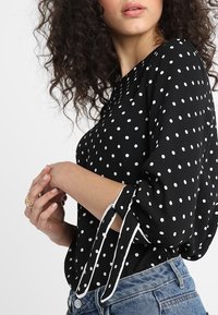 Černá halenka s bílými puntíky, s nabíranými rukávy, mašlí na manžetě a lehkou tkaninou. Nošená s vysokým pasem džín.