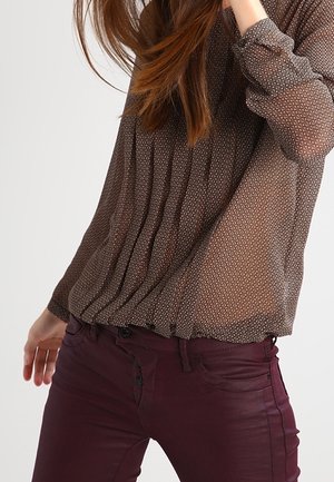 Frau trägt eine transparente, gemusterte braune Bluse und eng anliegende dunkelburgunderfarbene Hose, mit offenem Haar und einer Hand in der Nähe ihres Gesichts erhoben.