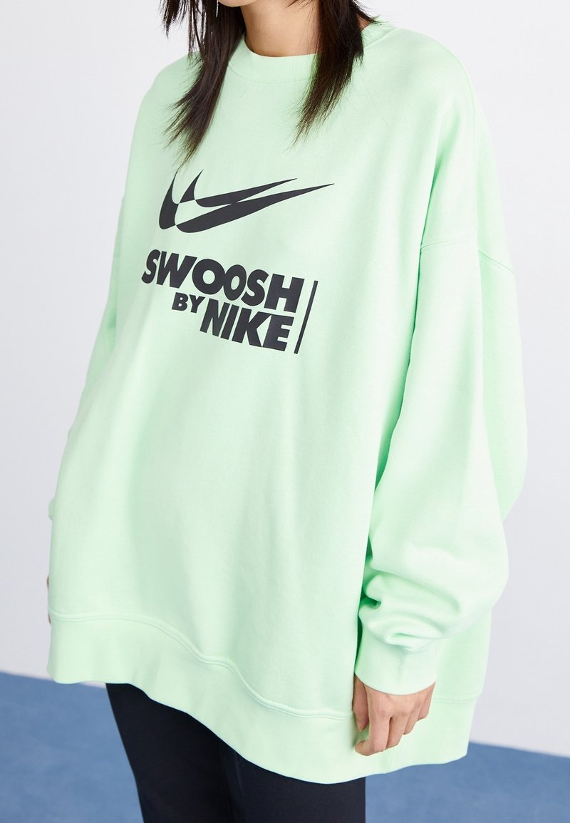 Sweatshirt oversize vert menthe avec manches longues, affichant un grand logo noir "Swoosh by Nike" à l'avant. Texture de tissu douce.