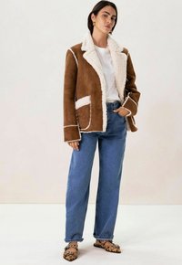 Veste en daim marron avec col et accents en shearling crème, associée à un pantalon large en denim bleu et des ballerines à imprimé léopard noir.