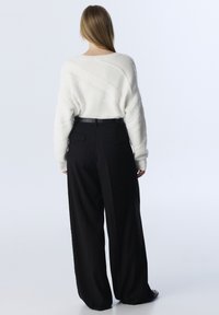 Žena z pohledu zezadu, oblečená v bílém strukturovaném svetru, černých kalhotách s širokými nohavicemi a páskem, a ve černých botách s hroty.