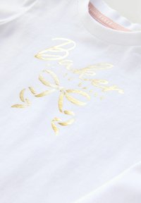 Biela košeľa so lesklým zlatým textom "Baker by Ted Baker" a dekoratívnym zlatým motýlikom pod textom.