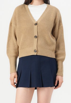 Femme portant un cardigan beige en maille épaisse avec trois boutons par-dessus un haut blanc et une mini-jupe plissée bleu marine.