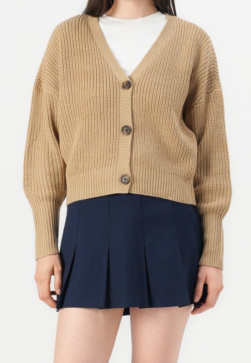 Femme portant un cardigan beige en maille épaisse avec trois boutons par-dessus un haut blanc et une mini-jupe plissée bleu marine.