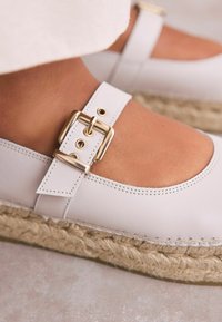 Weiße Leder-ESPADRILLES mit Cut-out-Design, verstellbarem Riemen und goldener Schnallen-Detail. Die Struktur der Jutewaren-Sohle ist hervorgehoben.