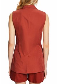 Bez rękawów marynarka w kolorze rdzy o gładkiej fakturze, z wysmuklającym kołnierzem i rozcięciem z tyłu. W komplecie z pasującymi szortami w tym samym odcieniu.