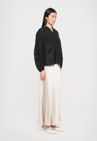 Sort langærmet top med knaplukning og teksturerede detaljer, parret med en let, beige satin maxi-nederdel og lyse slip-on sko.