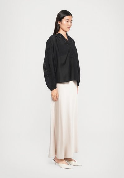 Top negru cu maneci lungi, cu un decolteu butonat și accente texturate, asortat cu o fustă maxi din satin bej, fluidă, și pantofi slip-on de culori deschise.