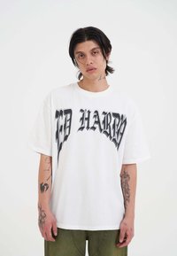 Witte oversized t-shirt met opvallende zwarte letters "ED HARDY" over de voorkant. De stof is glad; de mouwen zijn kort met een ontspannen pasvorm.