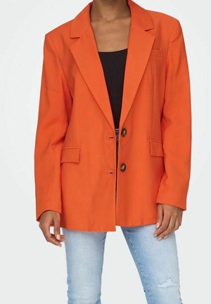 Femme portant un blazer orange vif à deux boutons sur un haut noir et un jean bleu clair déchiré, debout devant un fond blanc.