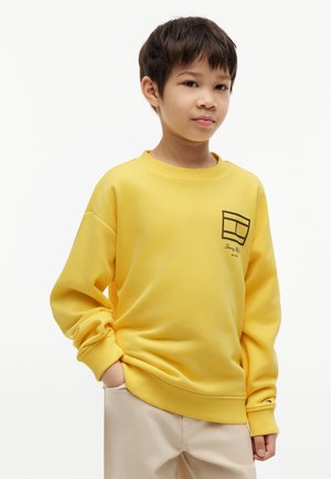 Boy standing with hands in pockets, wearing a bright yellow sweatshirt with a small graphic on the chest and beige pants.