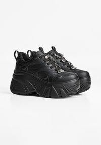 Een paar stoere zwarte platform sneakers met dikke gestructureerde zolen en zilveren kettingbedeltjes aan de veters tegen een witte achtergrond.