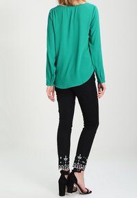 Blusa verde de manga larga con dobladillo redondeado, combinada con pantalones negros con bordados blancos en los puños y zapatos de tacón alto.