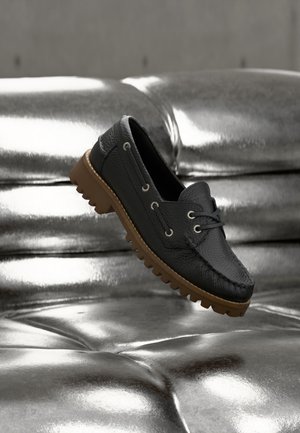 Mocasín de cuero negro con suela gruesa de goma marrón flotando sobre una superficie plateada brillante y acolchada.