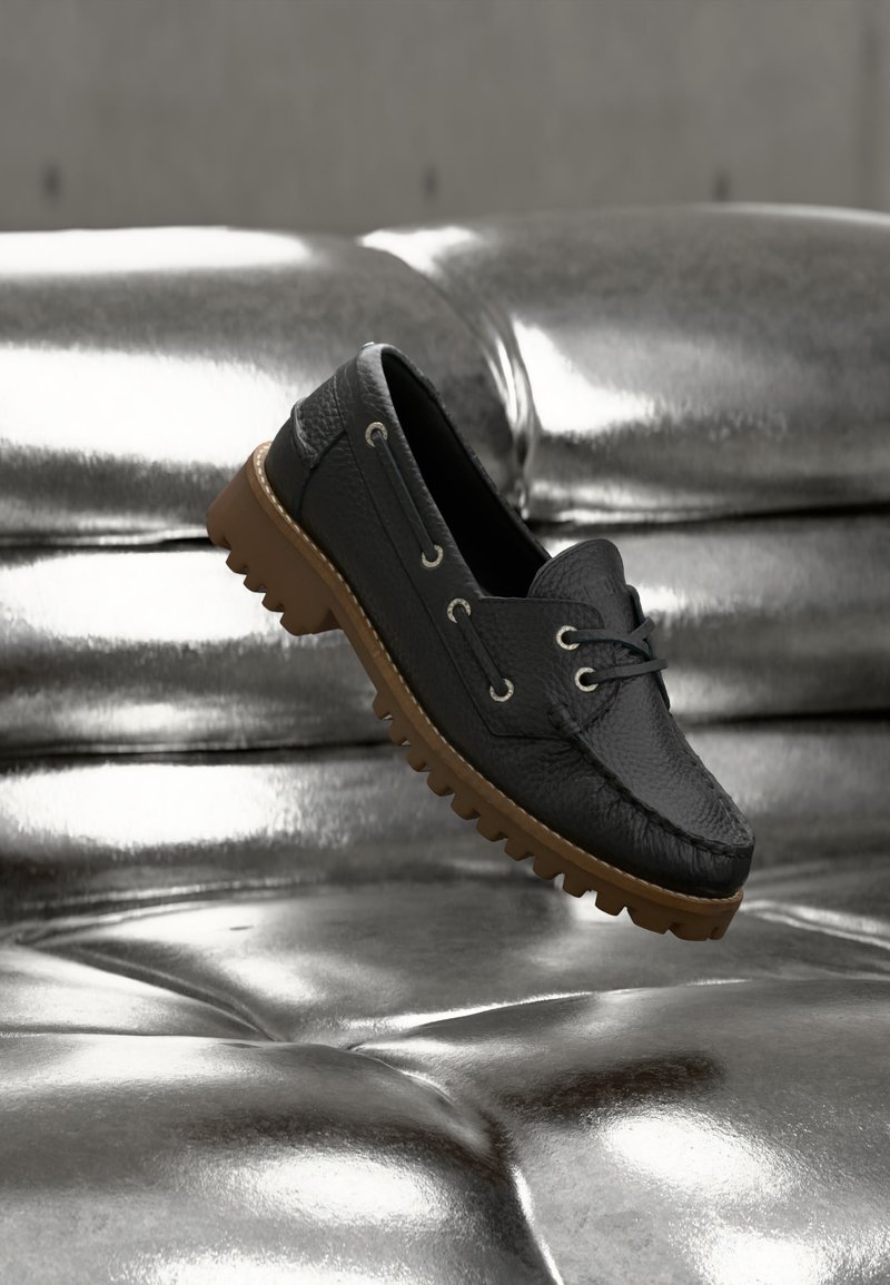 Mocassin en cuir noir avec une semelle épaisse en caoutchouc marron flottant au-dessus d'une surface argentée brillante matelassée.