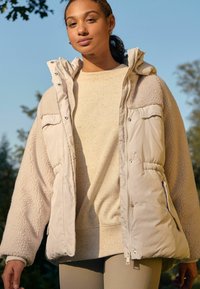 Veste rembourrée beige avec des manches douces, semblables à du polaire, des poches avant et une capuche. Portée sur un pull texturé beige clair et des leggings.
