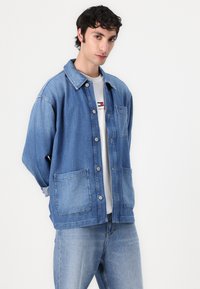 Lys blå denimjakke med knapping, to framlommer og spiss krage. Brukt over en hvit t-skjorte.