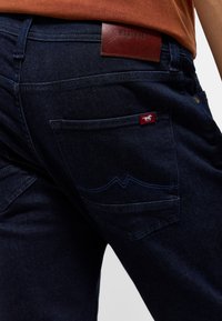 Ciemnoniebieskie dżinsy mają tylną kieszeń z czerwoną metką oraz skórzaną naszywkę w pasie. Wyraziste przeszycia podkreślają kieszenie.