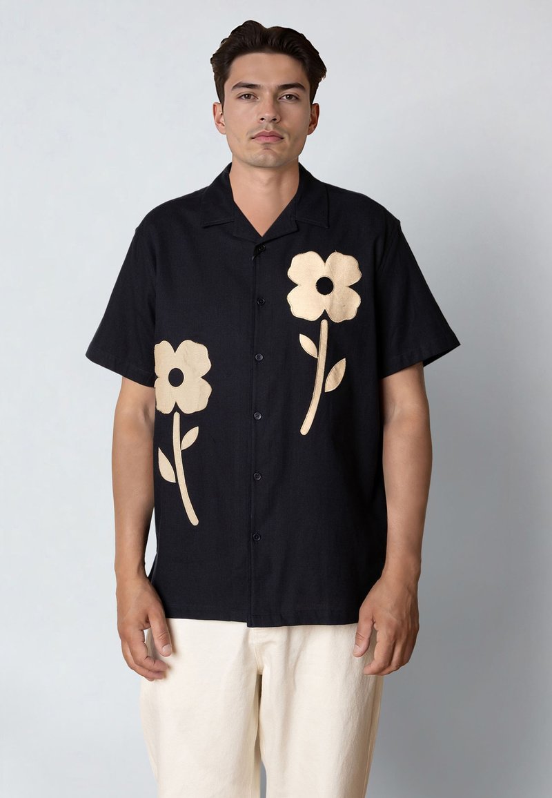 Homme portant une chemise noire à manches courtes avec de grands motifs de fleurs beiges sur le devant et un pantalon de couleur claire, sur un fond uni.