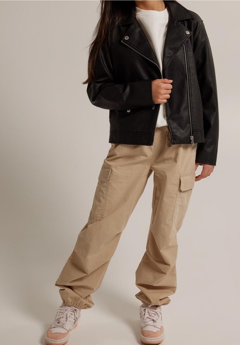 Giacca da moto in ecopelle nera con hardware argentato, chiusura con zip e colletto a punta, abbinata a pantaloni cargo beige e sneakers.