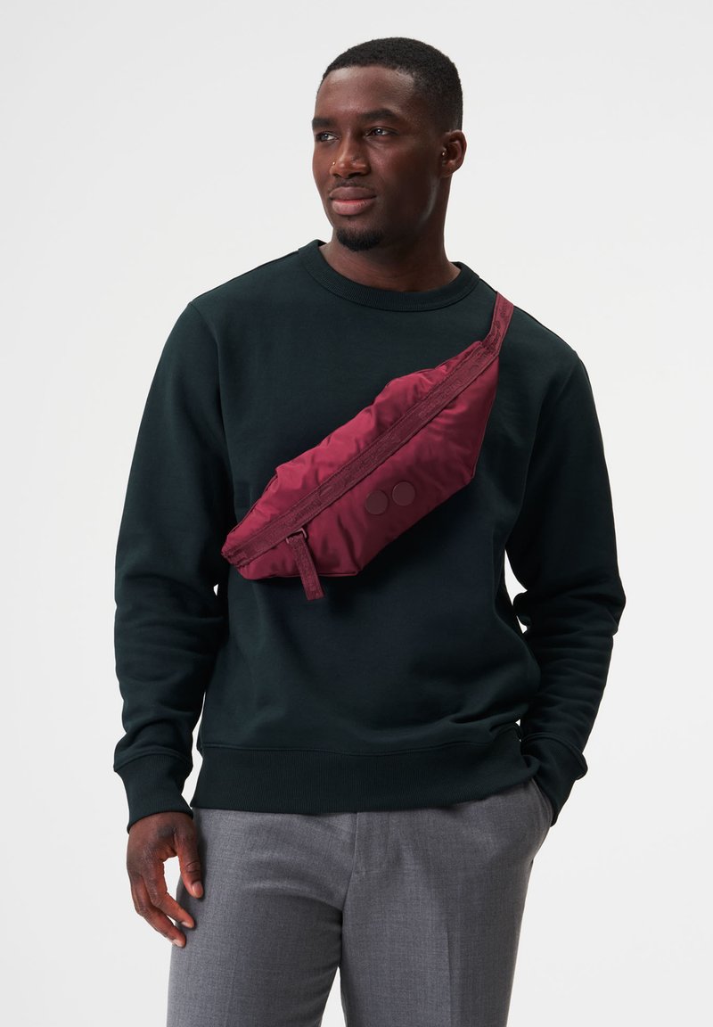 Bordeauxfarbene Umhängetasche aus weichem, gestepptem Stoff. Verfügt über einen Reißverschluss und zwei runde Akzente. Wird über einem schwarzen Sweatshirt getragen.