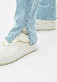 Valkoiset nahkakengät, joissa on rei'itettyjä yksityiskohtia ja kultaketjuakcentti, yhdistettynä vaaleansinisiin farkkuihin, joissa on sivurei'ät helman kohdalla.