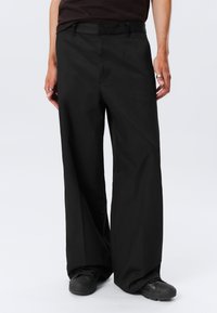 Pantalones de pierna ancha negros con una textura suave y presillas para cinturón. Presentan bolsillos frontales y un pliegue en la parte delantera, combinados con zapatos negros.