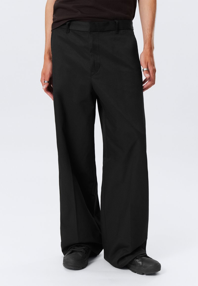 Pantalones de pierna ancha negros con una textura suave y presillas para cinturón. Presentan bolsillos frontales y un pliegue en la parte delantera, combinados con zapatos negros.