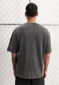 Grå bomulds t-shirt med korte ærmer, rund hals og afslappet pasform. Har et blåt logo på venstre ærme. Tekstureret stof med en falmet finish.