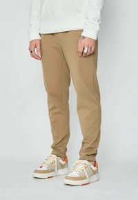 Personne portant un pantalon beige et des baskets blanches avec des accents orange, se tenant contre un fond gris clair uniforme.