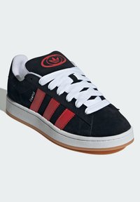 Sneaker in suede nero con accenti rossi, lacci bianchi e suola in gomma. Presenta tre strisce rosse e un logo circolare sulla linguetta.