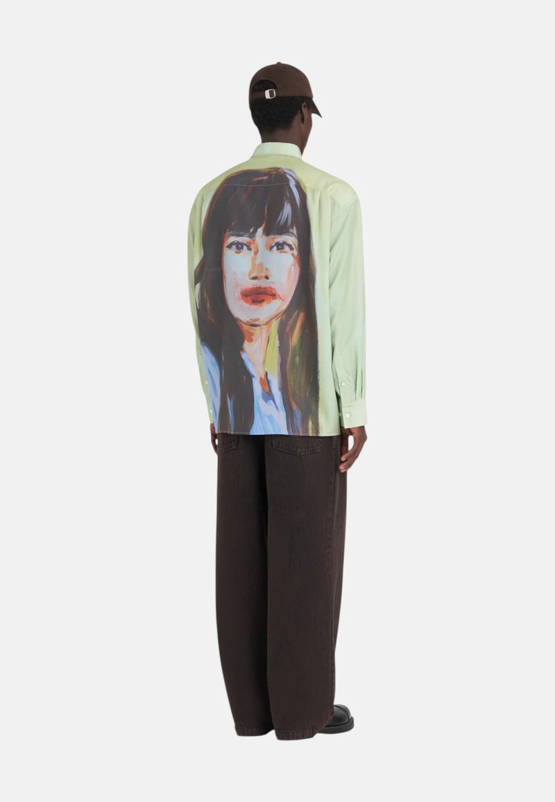 Chemise vert pastel avec une grande impression artistique de portrait au dos, présentant des couleurs vives. Associée à un pantalon large marron.