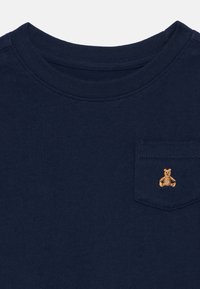 Mørkeblå kortermet T-skjorte i bomull, med en brystlomme som har et brodert teddy bear-design. Myk tekstur, rund hals.