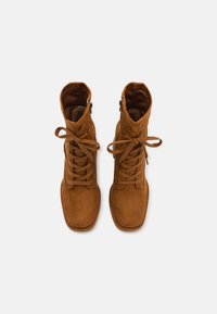 Bruna mockaskinnsankleboots med snörning framtill, fyrkantig tå och siddragkedja. Mjuk textur utan synliga mönster eller detaljer.