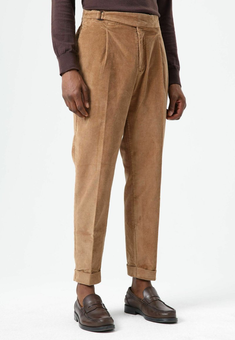 Brown corduroy trousers with a high waist, pleats, and rolled cuffs. Paired with dark brown loafers, highlighting a textured fabric.