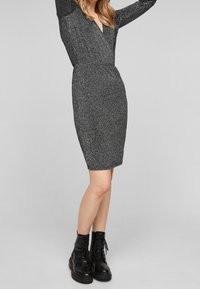 Svart glittrig stickad klänning med V-halsning och långa ärmar, figurnära midja och pen Silhouette; ihop med svarta snörda kängor.