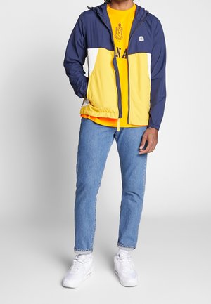 Chaqueta con bloques de color que presenta paneles en azul marino, amarillo y blanco. Tejido liviano, cierre con cremallera, capucha ajustable, combinada con una camiseta naranja y vaqueros.