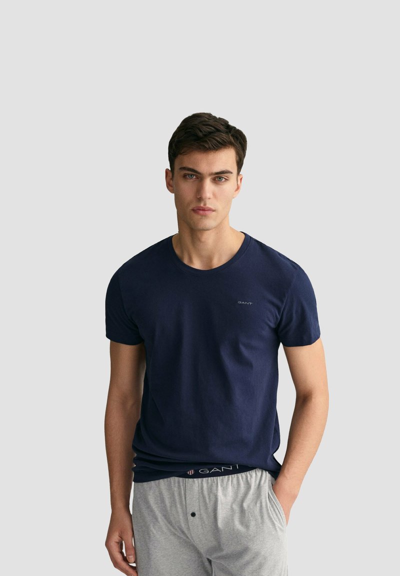 Giovane uomo con capelli scuri che indossa una maglietta GANT blu navy e pantaloni grigi, in piedi davanti a uno sfondo grigio chiaro uniforme.