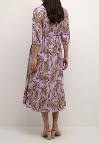 Fialové šaty s paisley vzorem v hnědých odstínech, se staženým pasem, tříčtvrtečními rukávy a vrstveným lemem. Nošené s béžovými sandály na bloku s podpatkem.