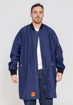 Mann trägt lange marineblaue Bomberjacke über weißem Hemd und Jeans und steht vor einfarbigem hellgrauem Hintergrund.