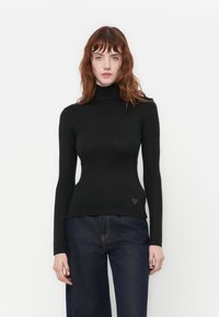Donna con capelli rossi che indossa un maglione a collo alto nero aderente e jeans blu scuri, in piedi contro uno sfondo bianco semplice.