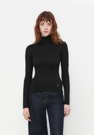 Donna con capelli rossi che indossa un maglione a collo alto nero aderente e jeans blu scuri, in piedi contro uno sfondo bianco semplice.