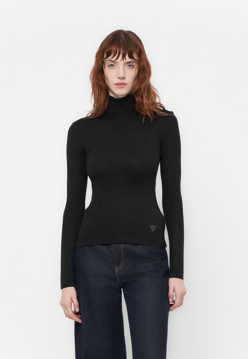Donna con capelli rossi che indossa un maglione a collo alto nero aderente e jeans blu scuri, in piedi contro uno sfondo bianco semplice.