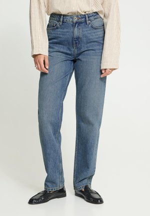 Femme portant un jean bleu droit, des mocassins noirs et une chemise beige à manches longues avec des rayures verticales, debout devant un fond uni.
