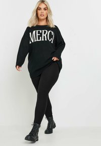 Pull en tricot noir avec "MERCI" en lettres blanches, associé à un legging noir côtelé et des bottines à lacets noires.
