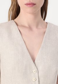 Beige sari vaateet, jossa on syvä V-pääntie ja kaksi beigeä nappia, esitellen teksturoitua kangasta ja muotoiltua istuvuutta.