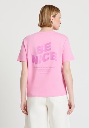 Persona con capelli biondi corti che indossa una maglietta rosa con la scritta "BE NICE" sul retro e pantaloni bianchi su uno sfondo semplice.