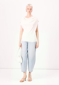 Ärmelloses helles Oberteil mit drapiertem Ausschnitt, hellblaue weite Jeans und beigefarbene Absatzsandalen. Das Model hält eine kleine weiße Handtasche.