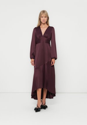 Largo vestido burdeos profundo con escote pronunciado, mangas abullonadas y dobladillo asimétrico. Tela de satén suave. Zapatos adornados con un lazo negro.