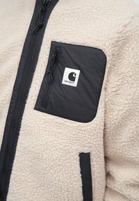 Villahuppari vaaleassa kreemissä, jossa on musta nailon-tasku, varustettuna vetoketjulla ja tekstuurilla. Taskussa on logopatch.